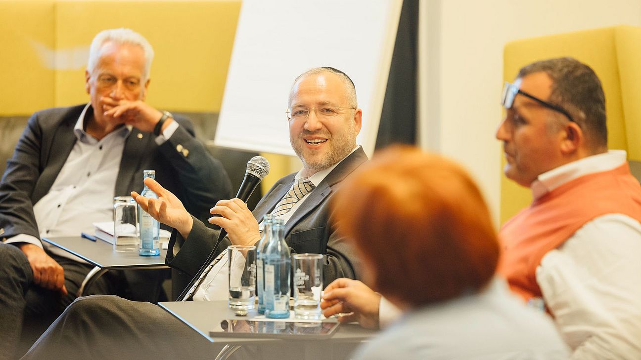 Vier Personen sitzen auf einem Podium, eine Person hält ein Mikrofon und spricht, auf Tischen stehen Wasserflaschen und Gläser.