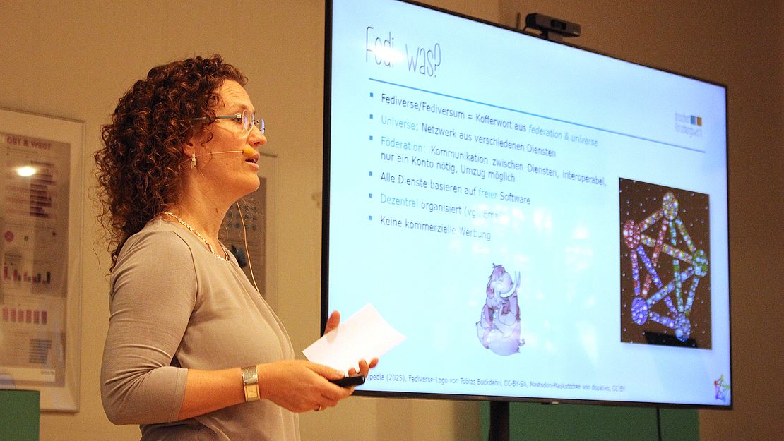 Frau mit lockigem Haar hält Notizen und präsentiert vor großem Bildschirm mit Text und Netzwerkgrafik zur Föderation von Diensten.
