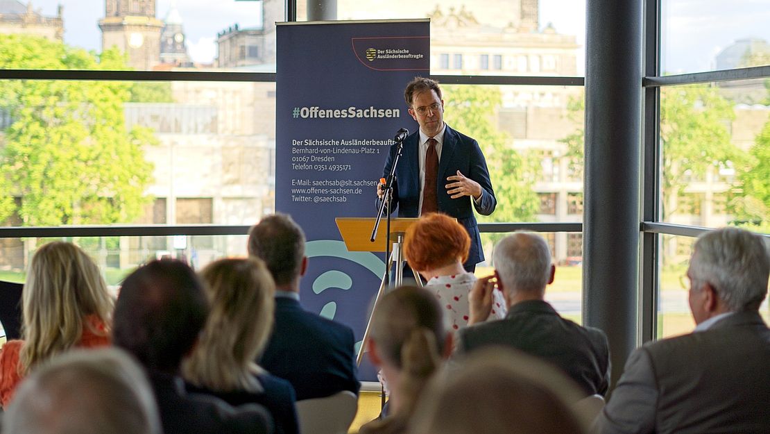 Prof. Dr. Johannes Eichenhofer spricht vor Publikum zum 20-jährigen Jubiläum der Sächsischen Härtefallkommission in einem modernen Veranstaltungsraum mit Blick auf Stadtgebäude.