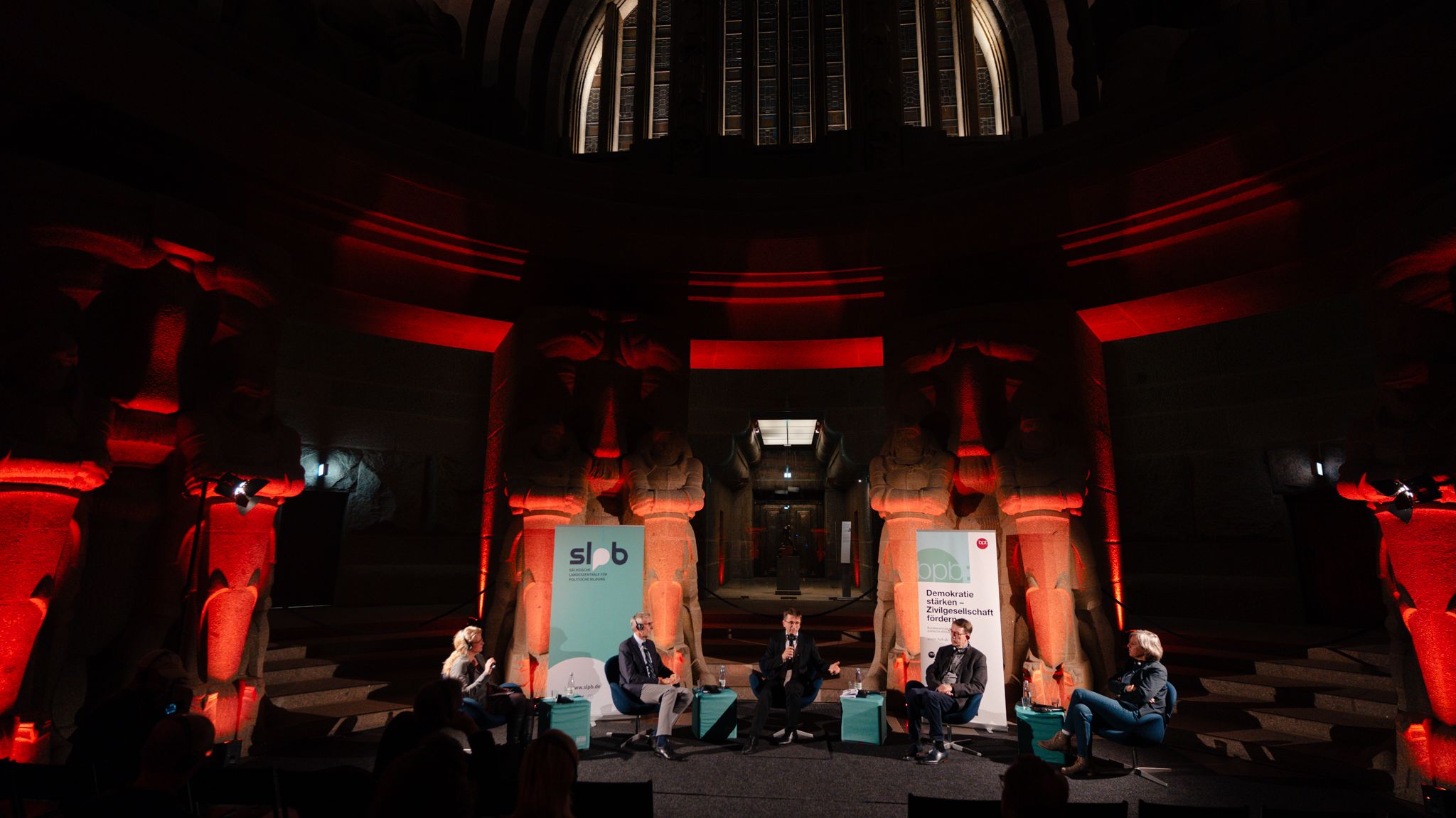 Fünf Personen sitzen auf einer Bühne im Leipziger Völkerschlachtdenkmal, umgeben von großen steinernen Skulpturen, rot beleuchtet, mit zwei Bannern im Hintergrund.