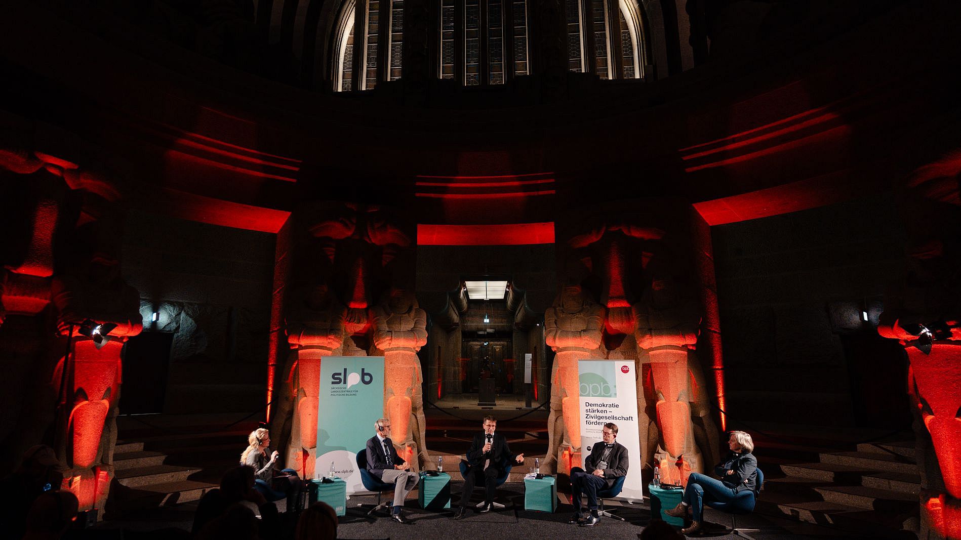 Fünf Personen sitzen auf einer Bühne im Leipziger Völkerschlachtdenkmal, umgeben von großen steinernen Skulpturen, rot beleuchtet, mit zwei Bannern im Hintergrund.