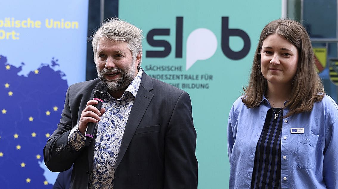 Zwei Personen stehen vor Bannern, eine hält ein Mikrofon, beide tragen Jacken, im Hintergrund Text 'Sächsische Landeszentrale für politische Bildung'.