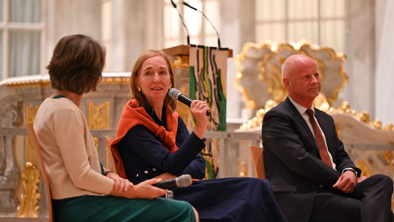 Drei Personen sitzen auf Stühlen in der Frauenkirche Dresden, eine Frau spricht in ein Mikrofon, im Hintergrund sind goldverzierte Kirchenbänke sichtbar.