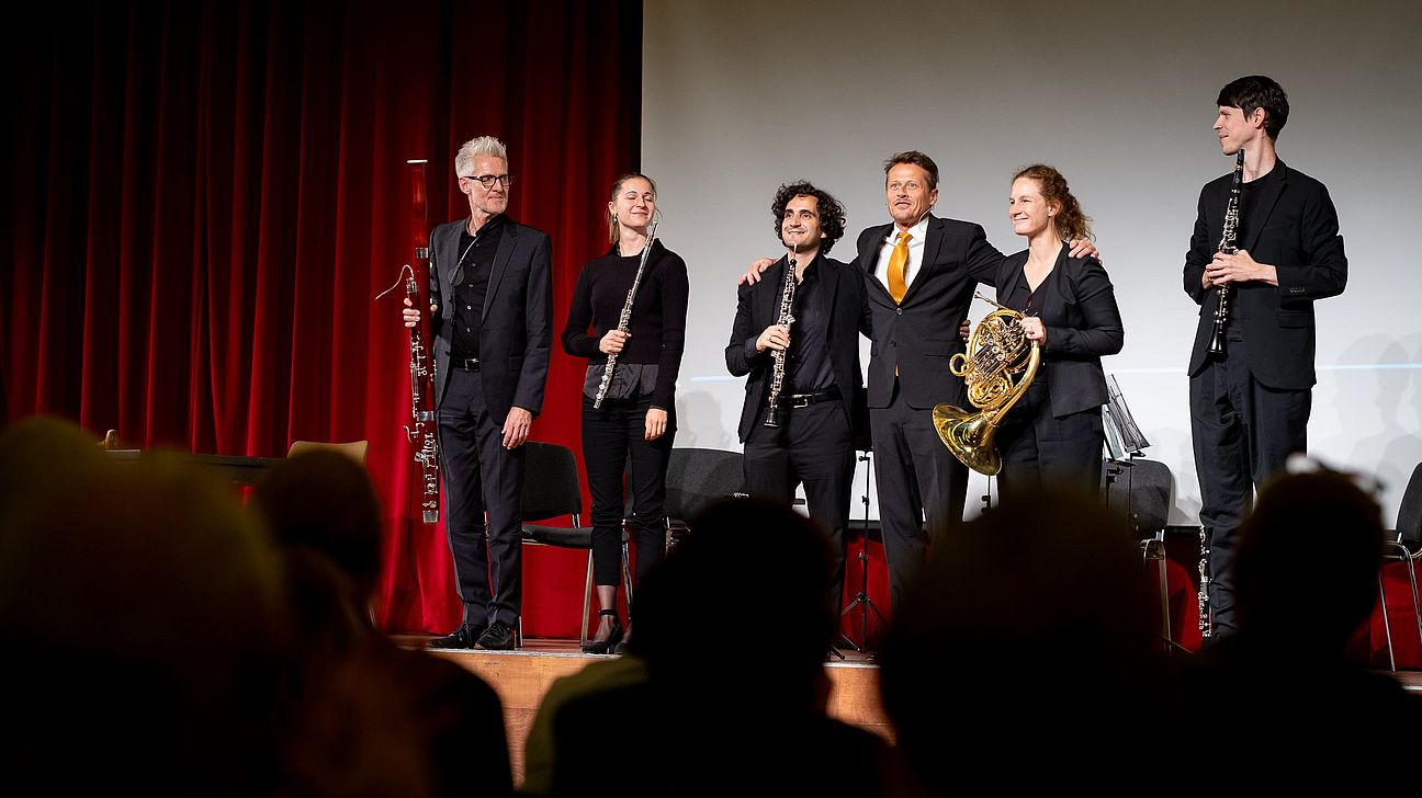 Sechs Musiker in schwarzer Kleidung stehen auf einer Bühne vor einem roten Vorhang, einige halten Blasinstrumente, darunter eine Klarinette und ein Horn.