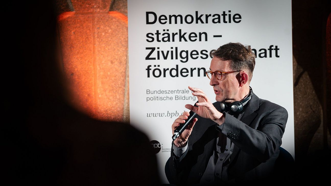 Winfried Kluth spricht mit Mikrofon vor einem Banner mit dem Text 'Demokratie stärken – Zivilgesellschaft fördern' im Leipziger Völkerschlachtdenkmal.