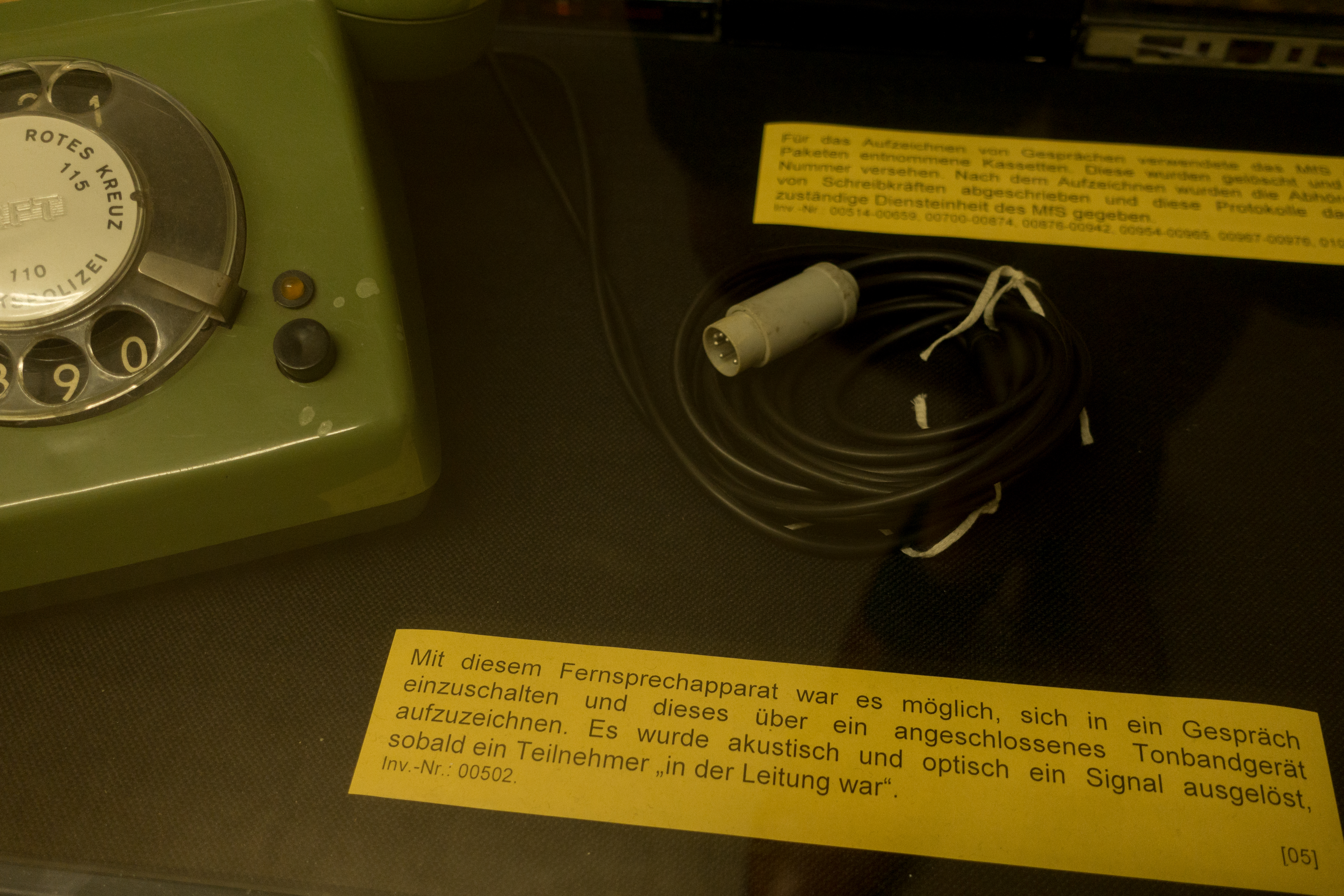 Das Bild zeigt ein historisches Überwachungsgerät. Es ist ein grünes Telefon mit Wählscheibe und einem angeschlossenen Kabel und Stecker. Daneben befindet sich ein gelbes Informationsschild mit dem Text: "Mit diesem Fernsprechapparat war es möglich, sich in ein Gespräch einzuschalten und dieses über ein angeschlossenes Tonbandgerät aufzuzeichnen. Es wurde akustisch und optisch ein Signal ausgelöst, sobald ein Teilnehmer 'in der Leitung war'." Es scheint, dass dies ein Gerät ist, das für Abhörzwecke verwendet wurde, wobei die genaue historische Epoche oder der Kontext des Gerätes nicht angegeben ist. Inv.-Nr.: 00502. Bei Klick vergrößert sich das Bild.