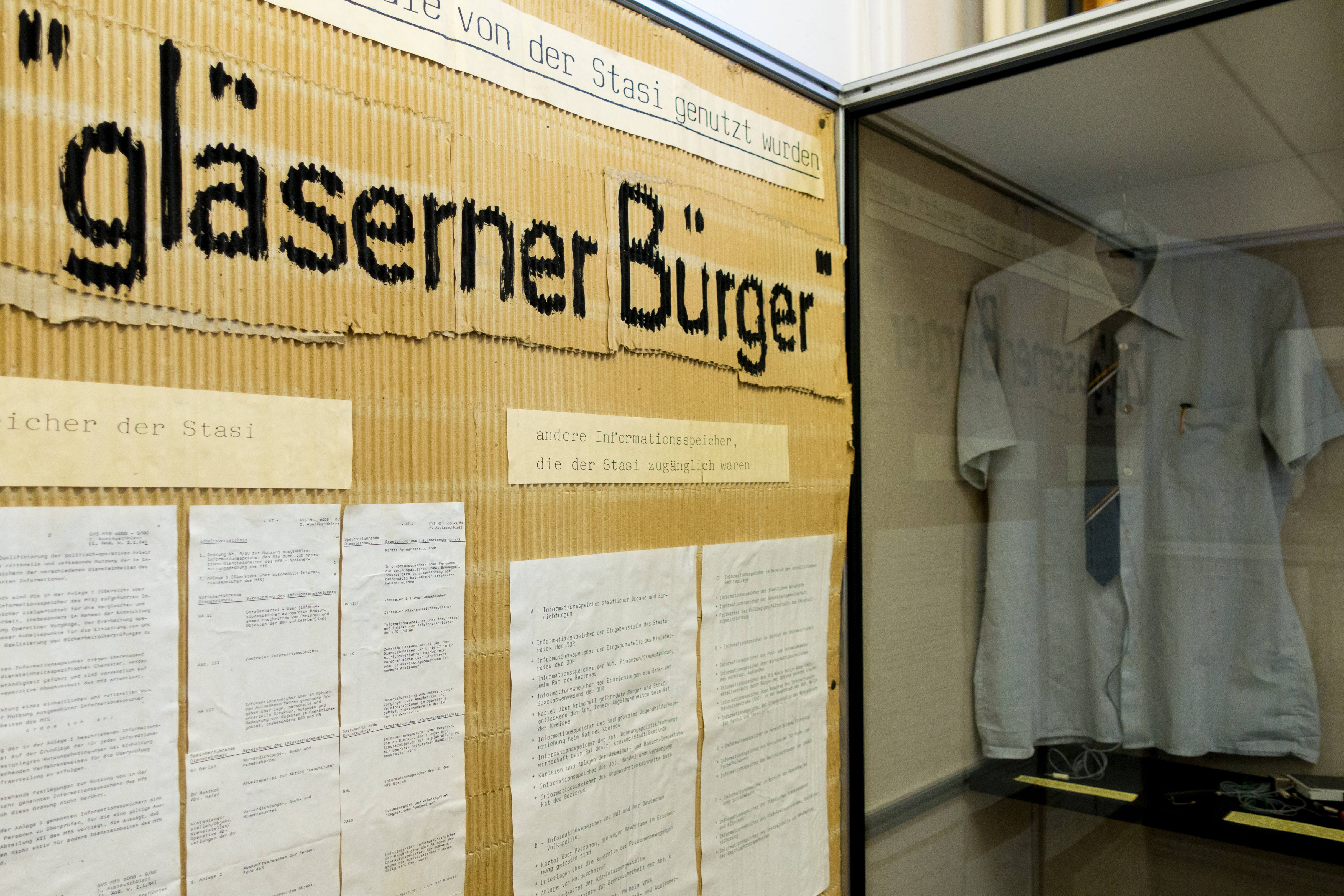 Das Bild zeigt eine Ausstellung mit Gegenständen und Dokumenten, die von der Stasi genutzt wurden. Im Vordergrund ist ein großes Schild mit der Aufschrift "gläserner Bürger" zu sehen, was sich auf die Überwachungsmethoden der Stasi bezieht, durch die Bürger transparent und ihre Privatsphäre durchsichtig gemacht wurden. Es sind verschiedene Papiere und Dokumente ausgestellt, die als "andere Informationsspeicher, die der Stasi zugänglich waren" beschrieben werden. Im Hintergrund ist eine Glasvitrine mit einem hellen Hemd zu erkennen, was möglicherweise Teil einer Uniform oder repräsentative Kleidung ist, die von Stasi-Mitarbeitern getragen wurde. Bei Klick vergrößert sich das Bild.