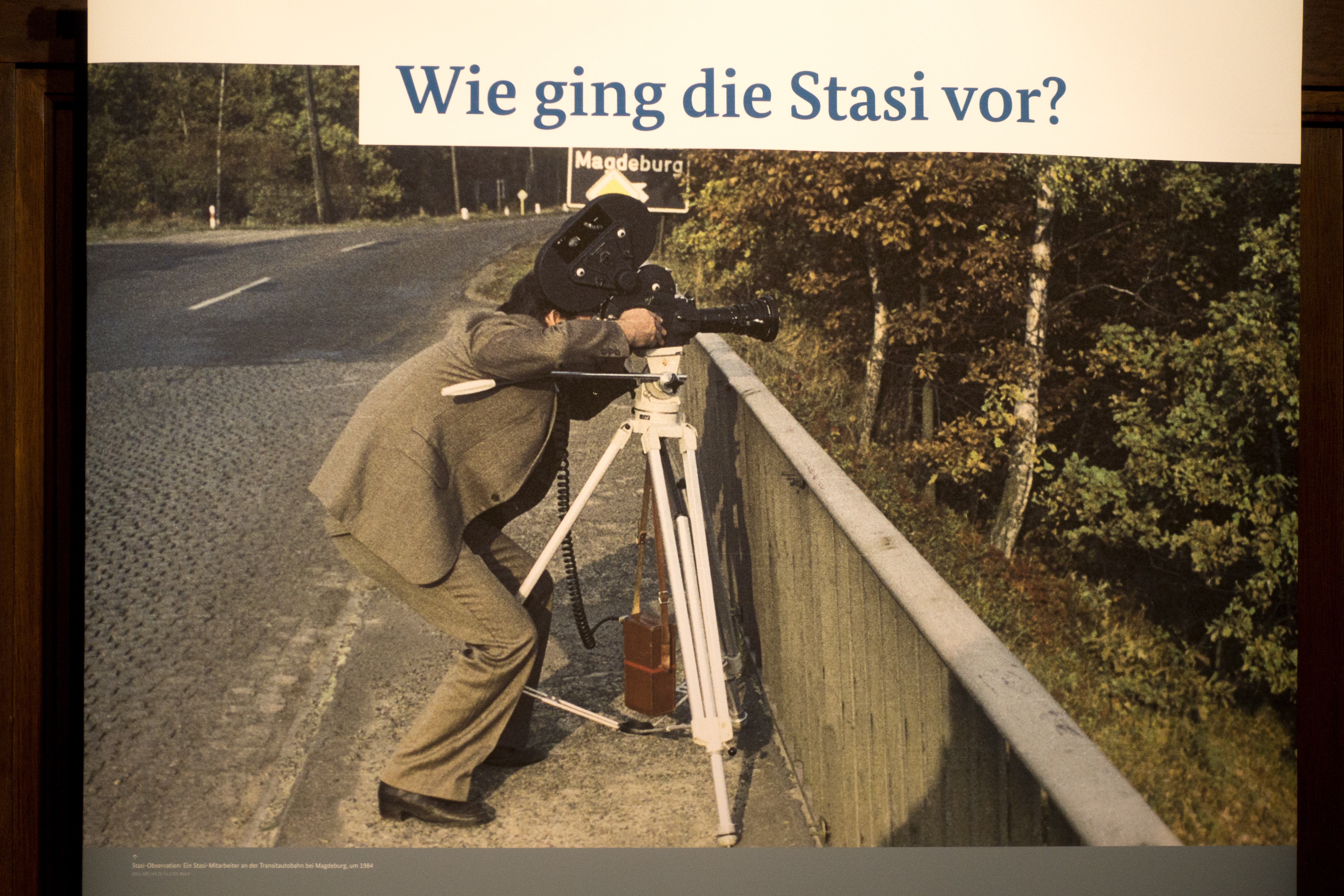 Das Bild zeigt ein Foto, das für eine Ausstellung verwendet wird und einen Stasi-Mitarbeiter im Jahr 1984 darstellt, wie er Überwachungsausrüstung bedient. Der Mitarbeiter ist in der Aktion zu sehen, wie er ein Fernglas oder ein anderes optisches Überwachungsinstrument auf einem Stativ bedient, das an einer Brücke aufgestellt ist. Im Hintergrund ist ein Straßenschild, das in Richtung Magdeburg zeigt. Über dem Foto steht der Text "Wie ging die Stasi vor?", was darauf hindeutet, dass die Ausstellung sich mit den Methoden und Vorgehensweisen der Staatssicherheit beschäftigt. Unter dem Bild befindet sich ein erläuternder Text, der allerdings im gegebenen Ausschnitt nicht vollständig lesbar ist. Bei Klick vergrößert sich das Bild.