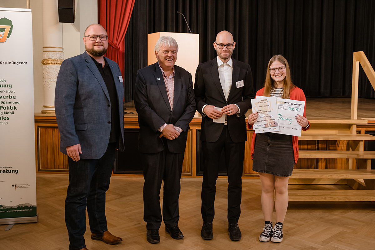 Sabine Bieberstein Auf dem Bild ist eine Preisverleihungsszene zu sehen. Drei Personen in Anzügen stehen neben einer jungen Person, die eine Urkunde und einen Scheck des "Jugend Engagement Wettbewerbs" hält. Der Scheck zeigt einen Betrag von 500 Euro. Im Hintergrund ist ein Banner mit der Aufschrift "Landeszentrale für politische Bildung" zu erkennen, was darauf hindeutet, dass die Veranstaltung einen politischen oder erzieherischen Kontext hat. Die Personen stehen auf einer Bühne, was durch die Holzstufen und den Vorhang im Hintergrund hervorgehoben wird.