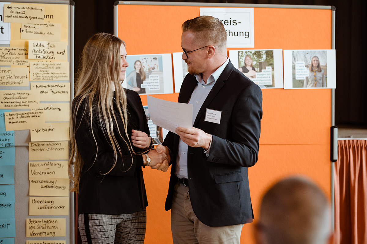 Fee Wurzener Land Auf dem Bild ist ein Handschlag zwischen zwei Personen zu sehen, die sich Dokumente übergeben. Im Hintergrund ist eine Pinnwand mit vielen Post-it-Notizen und ein Plakat mit der Aufschrift "Preisverleihung". Die Post-it-Notizen scheinen Kommentare oder Feedback zu beinhalten, was auf eine interaktive Sitzung oder einen Workshop hinweisen könnte. Beide Personen sind halb von der Seite fotografiert, wobei der Fokus auf der Übergabe der Dokumente und dem Handschlag liegt.
