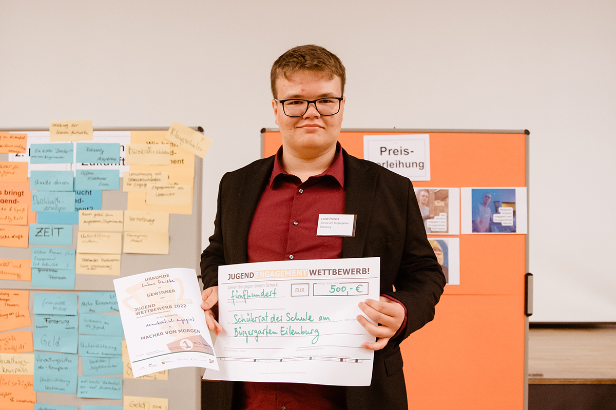 Lukas Eilenburg Das Bild zeigt eine Person, die stolz eine Urkunde und einen symbolischen Scheck des "Jugend Engagement Wettbewerbs" präsentiert. Die Urkunde trägt den Titel "Gewinner" und ist für den Wettbewerb 2022 ausgezeichnet worden, unter dem Motto "MACHER VON MORGEN". Der Scheck zeigt einen Betrag von 500 Euro und ist ausgestellt für den "Schülerrat der Schule am Bürgergarten Eilenburg". Im Hintergrund sind Post-it-Notizen und ein Poster zur "Preisverleihung" zu erkennen, was darauf hindeutet, dass die Szene in einem Workshop- oder Konferenzraum aufgenommen wurde.