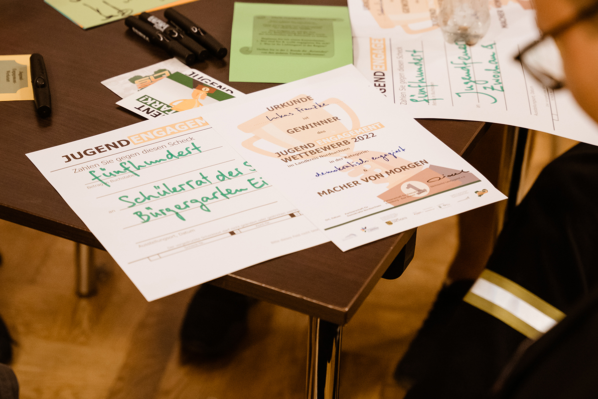Lukas Eilenburg Das Bild zeigt einen Tisch, auf dem verschiedene Dokumente ausgebreitet sind, darunter eine Urkunde und ein symbolischer Scheck des "Jugend Engagement Wettbewerbs". Die Urkunde hat die Überschrift "Gewinner" und ist für den Wettbewerb 2022 ausgestellt. Auf dem Scheck ist der Name "Schülerrat des Bürgergarten Ei" zu lesen, was vermutlich auf den "Schülerrat des Bürgergartens Eilenburg" hinweist, und der Betrag ist mit "Fünfhundert" angegeben, also 500 Euro. Neben den Dokumenten liegen auch Schreibutensilien auf dem Tisch, was darauf schließen lässt, dass die Dokumente möglicherweise gerade unterzeichnet wurden oder zur Unterschrift bereit liegen.