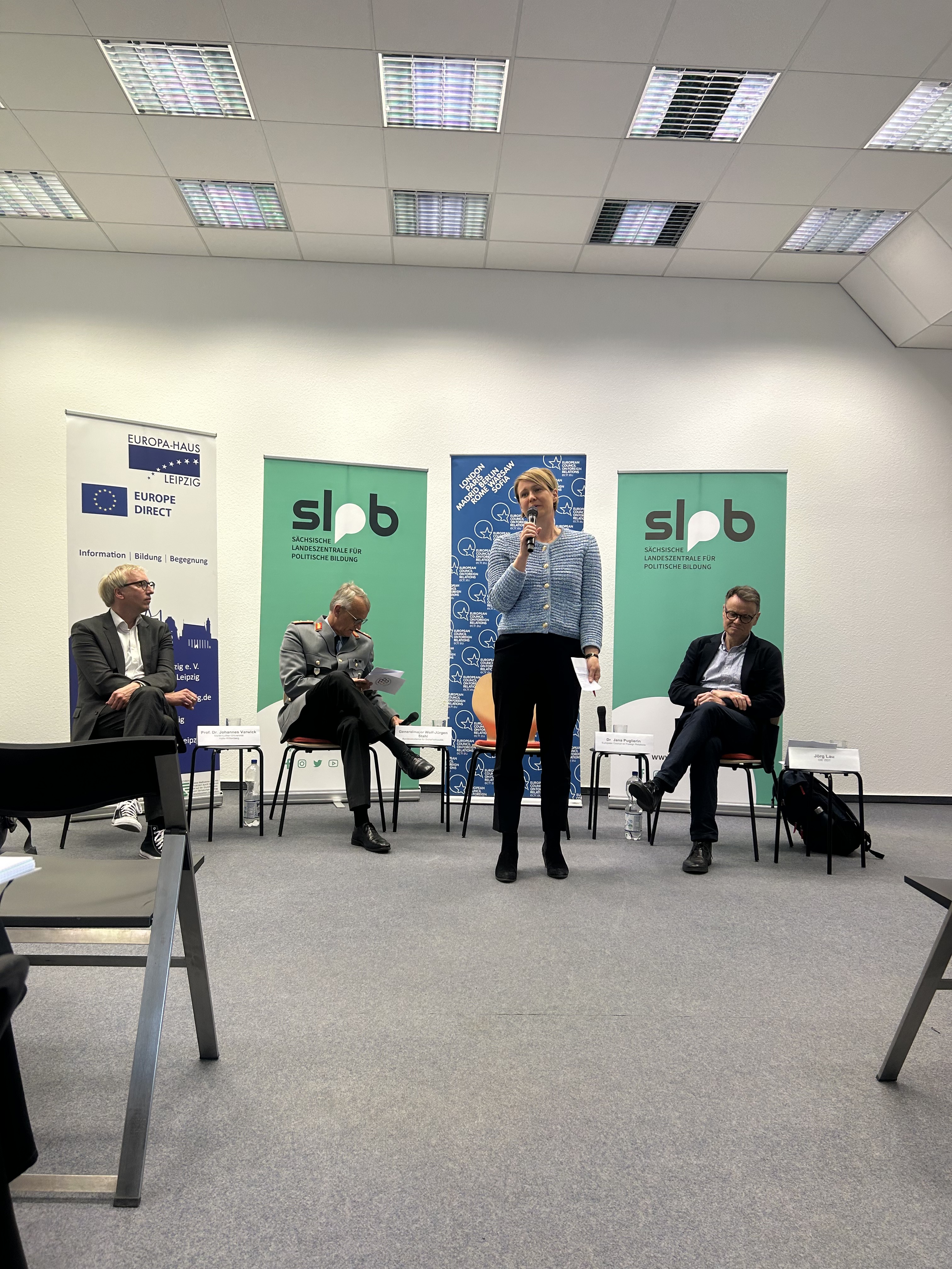 Vor Roll Ups sitzen drei Menschen. Zwischen ihnen steht Jana Puglierin, Leiterin des ECFR mit einem Mikrofon in der Hand