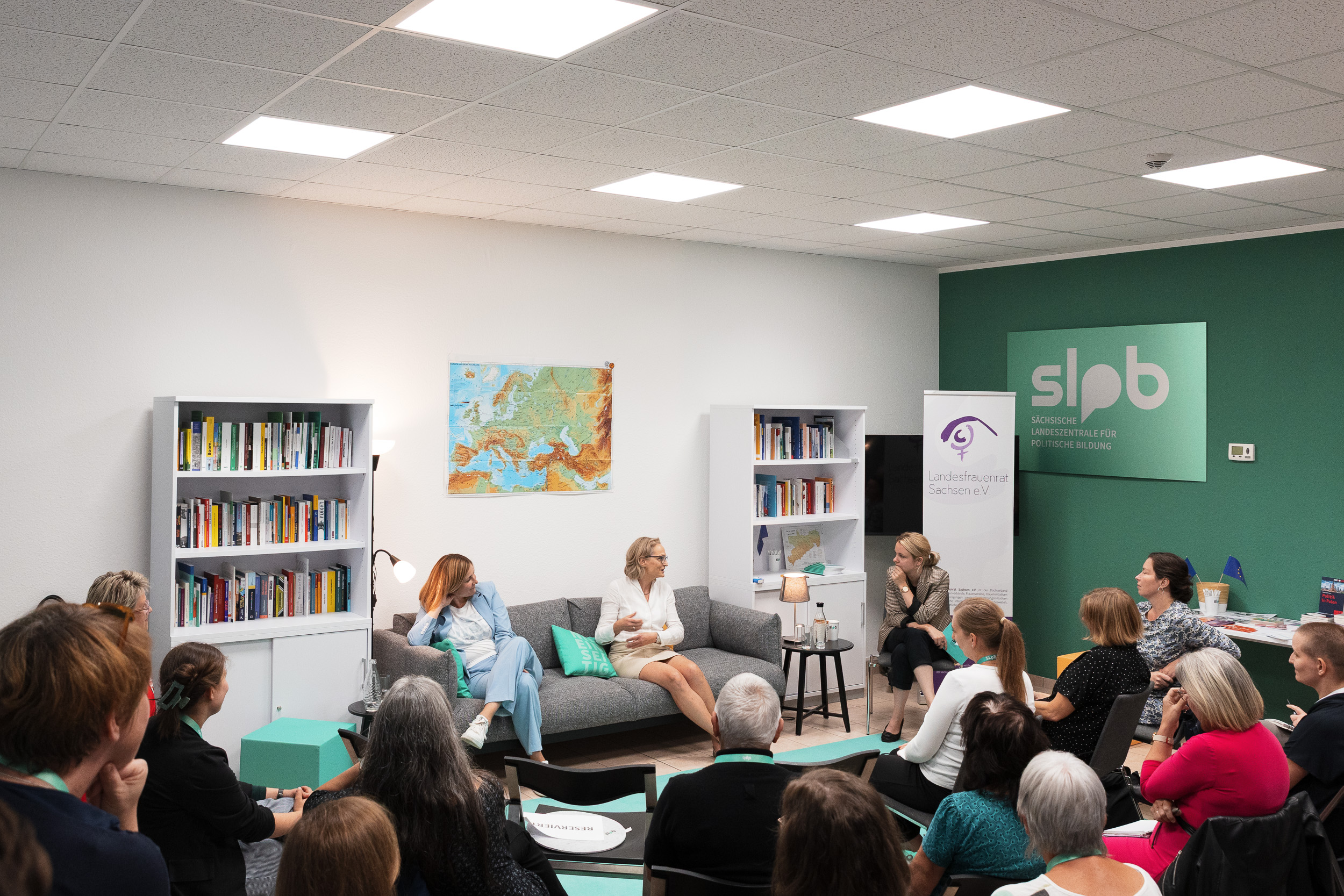 Zwei Frauen sitzen auf einem grauen Sofa in einem Raum mit Bücherregalen. Eine Frau hält ein Mikrofon, während die andere spricht. Im Hintergrund steht ein Schild mit 'slpb'.