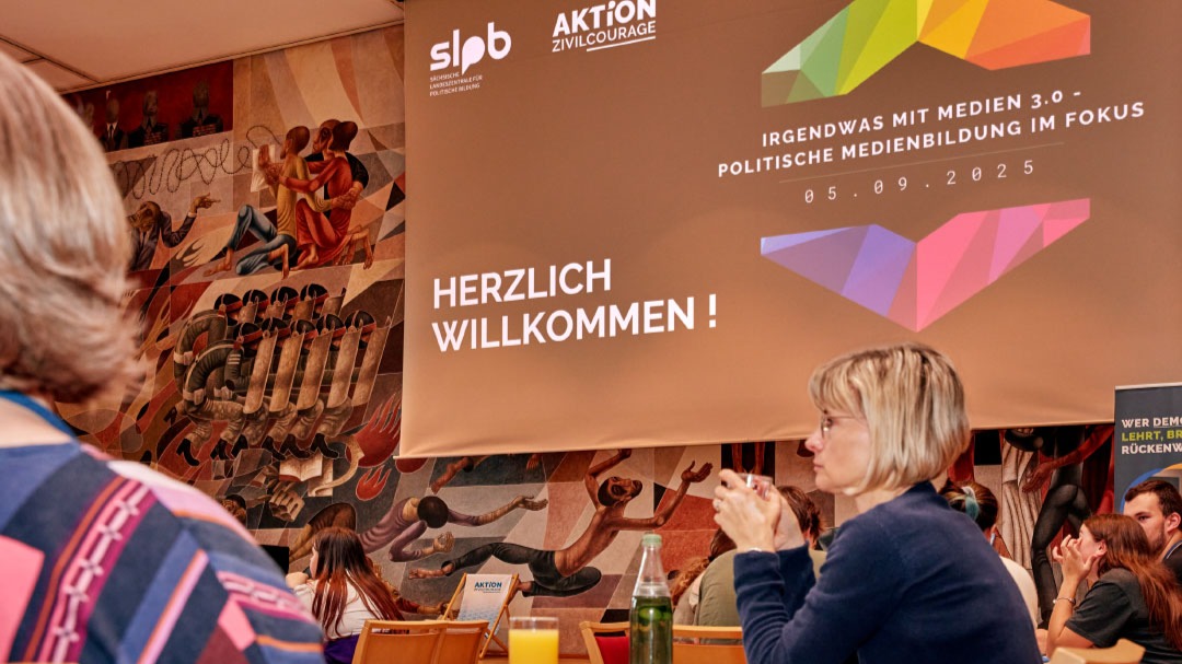 Zwei Personen von hinten in einem Raum mit einer Wandmalerei. Ein Banner zeigt 'HERZLICH WILLKOMMEN !' und 'IRGENDWAS MIT MEDIEN 3.0 - POLITISCHE MEDIENBILDUNG IM FOKUS 05.09.2025'.