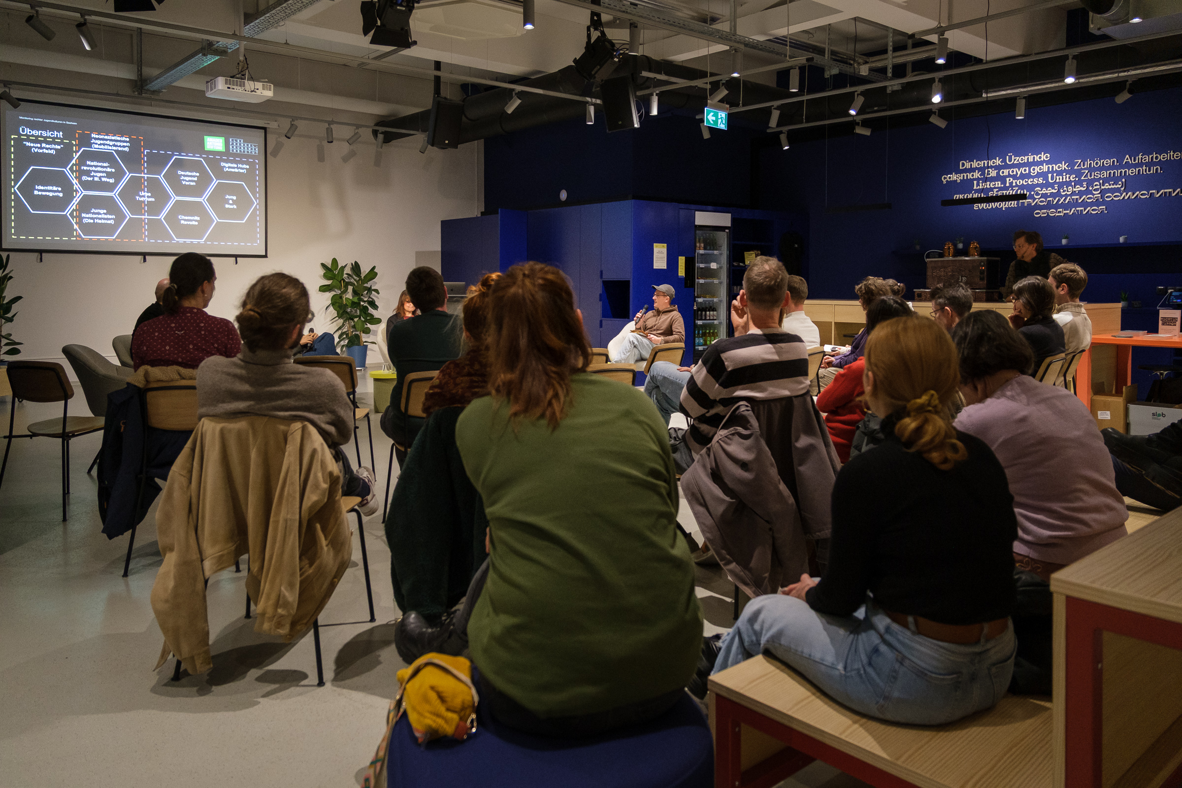 Mehrere Personen sitzen in einem Raum und schauen auf eine Präsentation mit Text und Diagrammen an der Wand.