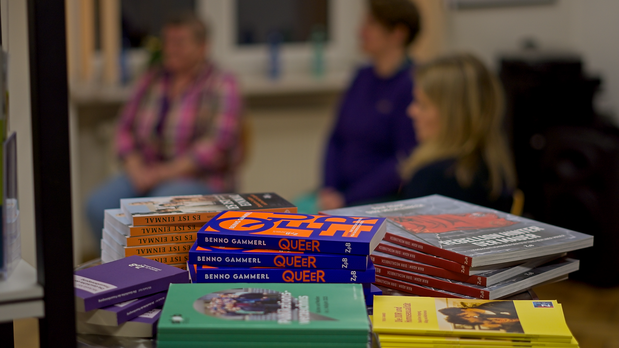 Im Hintergrund sind die drei Podiumsgäste unscharf zu erkennen. von links: Elke Prinz, Barbara Wallbraun und Teresa Tammer. Im Vordergrund ist scharf ein Büchertisch zu erkennen.