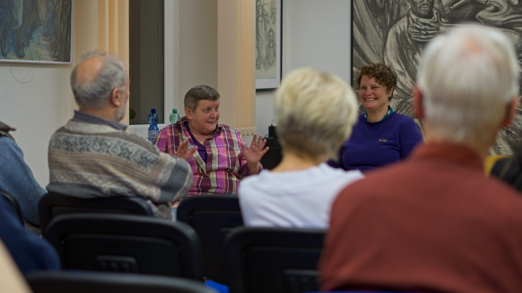 Elke Prinz und Barbara Wallbraun sind im Hintergrund zu sehen, wie sie vom Podium zu den anwesenden Gästen sprechen.