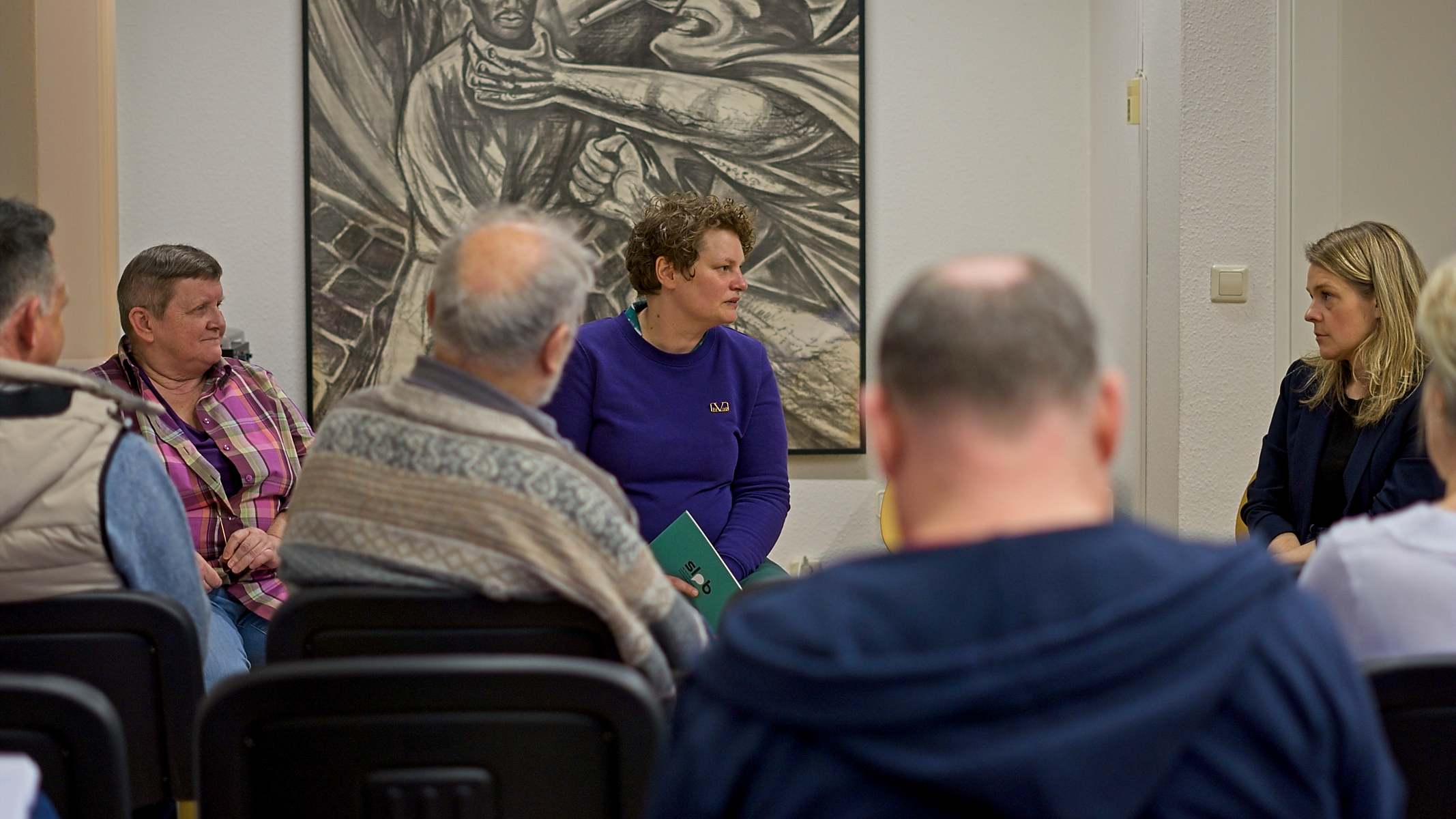 Im Hintergrund ist das Podium zu erkennen, dass vor einem großen Gemälde sitzt. von links: Elke Prinz, Barbara Wallbraun und Teresa Tammer. Im Vordergrund sitzen mehrere Gäste, die unscharf sind.